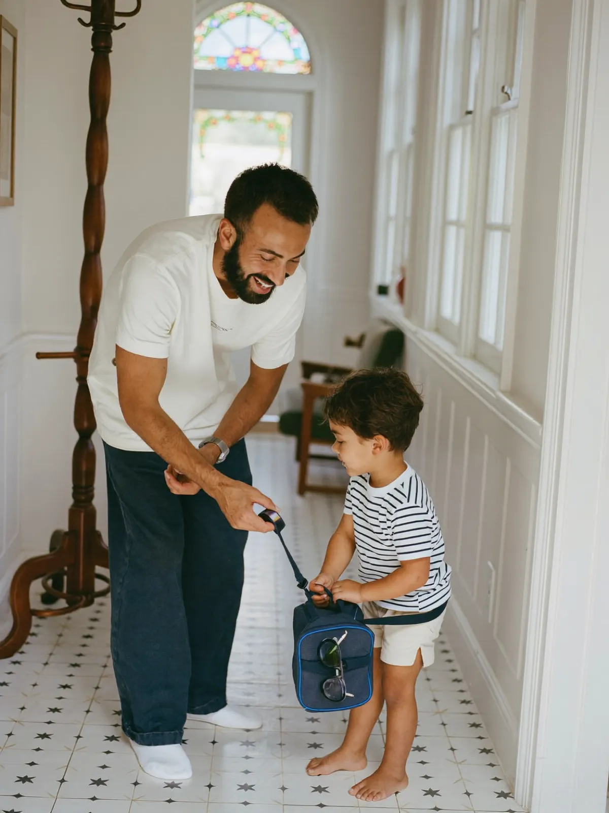 A dad crouches in the hallway to help his young son with his swimming bag, both smiling.